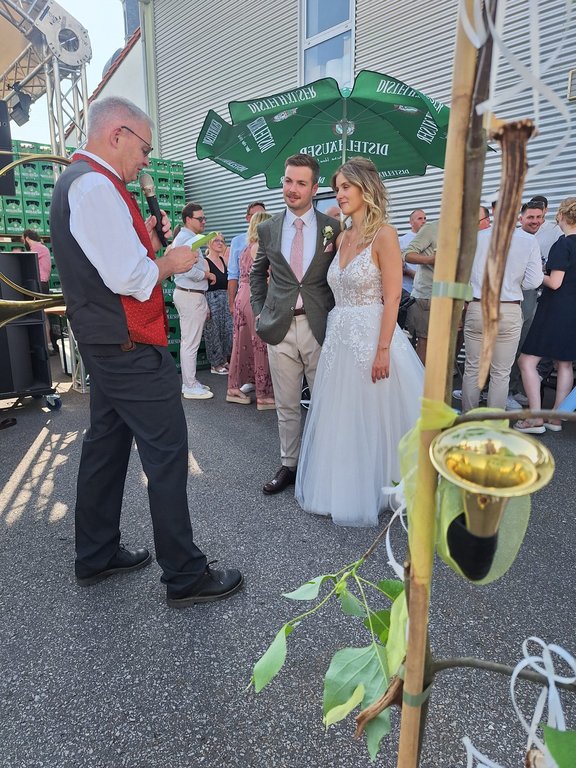Ein Brautpaar steht nebeneinander, während ein Mann mit Mikrofon spricht; im Hintergrund sind Gäste, Getränkekisten und ein grüner Sonnenschirm mit der Aufschrift "Pilsner Urquell" zu sehen.