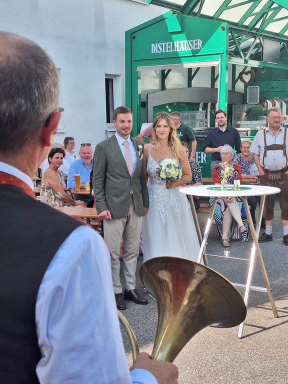 Brautpaar steht lächelnd vor einem grünen Distelhäuser-Stand, umgeben von Gästen, während eine Person mit einem Blasinstrument spielt. Im Hintergrund sitzen Menschen an Biertischen.