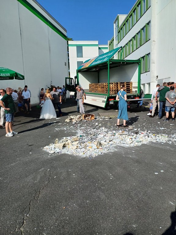 Ein LKW mit offener Ladefläche steht auf einem Hof zwischen Gebäuden. Menschen in alltäglicher und festlicher Kleidung umringen einen großen Haufen von zerbrochenem Porzellan auf dem Boden.