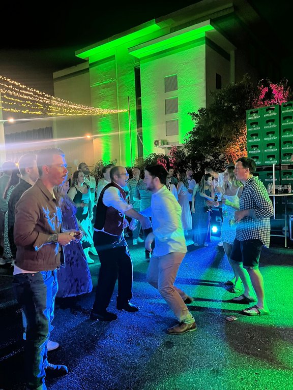 Personen tanzen auf einer nächtlichen Party im Freien vor einem grünlich beleuchteten Gebäude. Über ihnen hängen Lichterketten, und im Hintergrund sind gestapelte Getränkekästen.