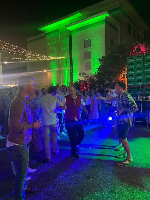 Personen tanzen ausgelassen auf einer bunt beleuchteten Party im Freien, mit einem Gebäude im Hintergrund. Lichterketten und Bierkisten sorgen für eine festliche Atmosphäre.