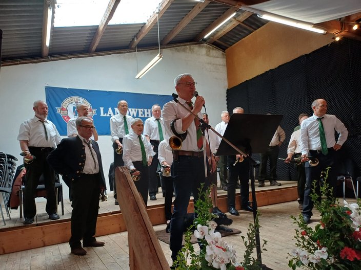 Eine Gruppe älterer Männer mit Hörnern steht auf einer Bühne. Einer spricht ins Mikrofon. Im Hintergrund hängt ein Banner mit der Aufschrift: "LANDWEHR." Der Raum ist dekoriert.