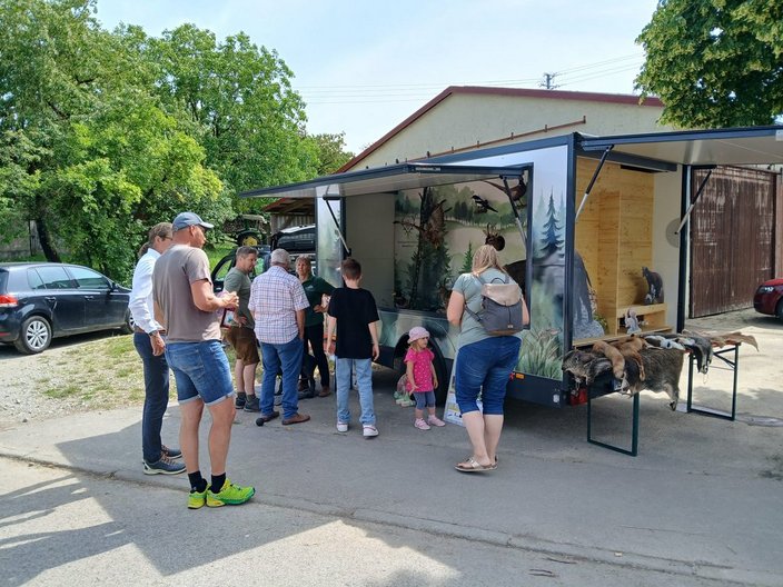 Anhänger – steht geöffnet – auf einem Hof, zeigt ausgestopfte Waldtiere; Menschen stehen drumherum und betrachten die Ausstellung. Links parkt ein Auto, Bäume im Hintergrund.