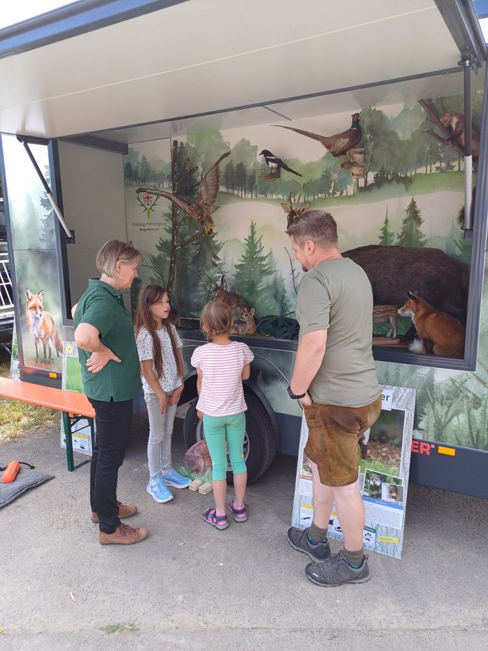 Ein mobiler Infostand zeigt ausgestopfte Tiere, umgeben von Menschen, die diskutieren. Im Hintergrund sind Waldmotive. Text: "Kreisjägervereinigung Bergheim e.V." und daneben Informationsplakate.
