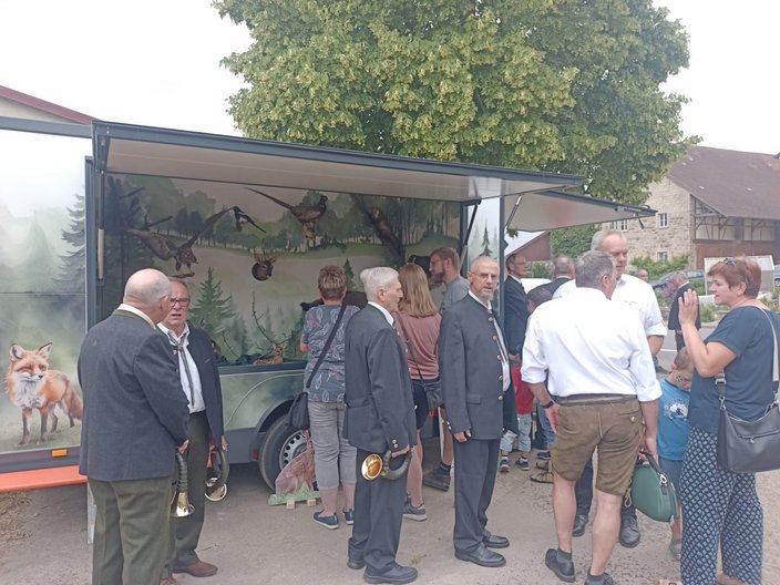 Anhänger zeigt Waldmotiv mit Tieren. Menschen stehen davor, plaudern oder betrachten die Szene. Bäume und Gebäude im Hintergrund schaffen eine ländliche Atmosphäre. Keine sichtbaren Texte.