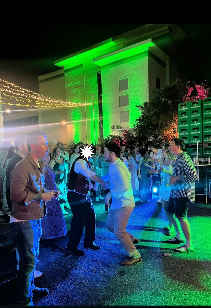 Personen tanzen beleuchtet auf einer nächtlichen Outdoor-Party. Im Hintergrund ist ein grünes, beleuchtetes Gebäude sichtbar, dekoriert mit Lichterketten. Rechts steht neben Pflanzen ein Getränkekastenstapel.