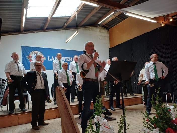 Ein Chor aus älteren Männern steht auf einer Bühne und spielt Jagdhörner. Sie tragen weiße Hemden und grüne Krawatten. Im Hintergrund hängt ein Banner mit der Aufschrift "LANDWEHR".
