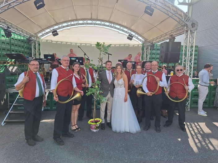 Musiker mit Hörnern posieren neben einem Paar, wahrscheinlich Brautpaar. Die Gruppe steht vor einer Bühne mit überdachtem Pavillon. Im Hintergrund sind Getränkekisten und weitere Gäste zu sehen.