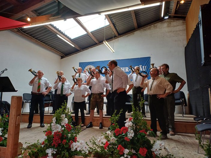 Männer und eine Frau spielen Hörner auf einer Bühne. Sie tragen formelle Kleidung. Im Hintergrund ist ein Banner mit „WURZNER – BRAU“ zu sehen. Blumen schmücken die Vorderseite der Bühne.