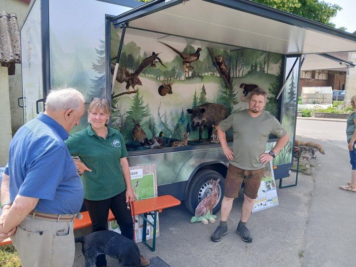 Ein mobiler Ausstellungsstand zeigt ausgestopfte Wildtiere in einem naturnahen Hintergrund. Eine Frau und ein Mann stehen daneben, in Gespräch mit einem älteren Herrn. Ein Hund schnuppert herum.